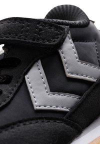 Sneaker nera con tracolla in velcro, caratterizzata da cuciture a zigzag grigie e dettagli, realizzata in morbido tessuto e materiale scamosciato. Finitura texturizzata.