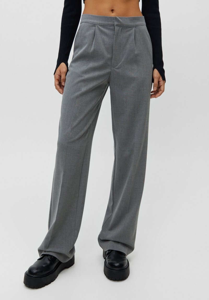 Femme portant un pantalon gris taille haute à jambes larges et des chaussures compensées noires, debout devant un fond clair uni.