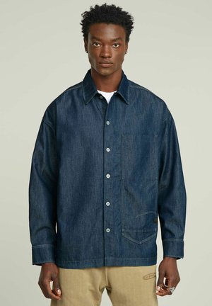 Mann trägt dunkelblaues Jeanshemd mit Knöpfen und großer Fronttasche sowie beige Hose, steht vor einfarbig hellem Hintergrund.