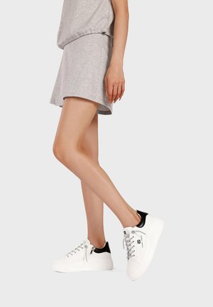 Donna con gonna e top grigio chiaro che indossa sneakers bianche platform con dettagli neri sul tacco, in piedi con un piede leggermente sollevato.