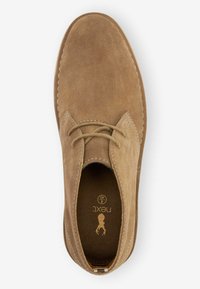Zapato de ante marrón con cordones y punta redondeada, costuras en contraste y un interior texturizado. Cuenta con un logo impreso en la plantilla.