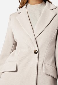 Blazer beige testurizzato con rever appuntiti, chiusura a un solo bottone e due tasche frontali. Il tessuto presenta un delicato motivo a righe.