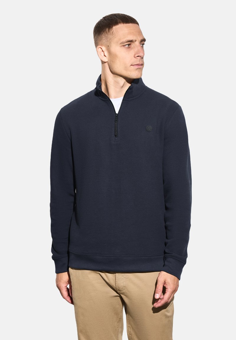 Muž oblečený v námořnickém modrém pulloveru na čtvrt zip a béžových kalhotách, dívající se doprava na bílém pozadí.