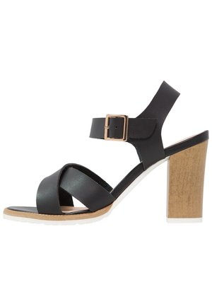Czarna sandałka na obcasie z paskami krzyżującymi się, klamrą przy kostce i masywnym obcasem o fakturze drewna. Gładka tekstura skóry i miękka wkładka.