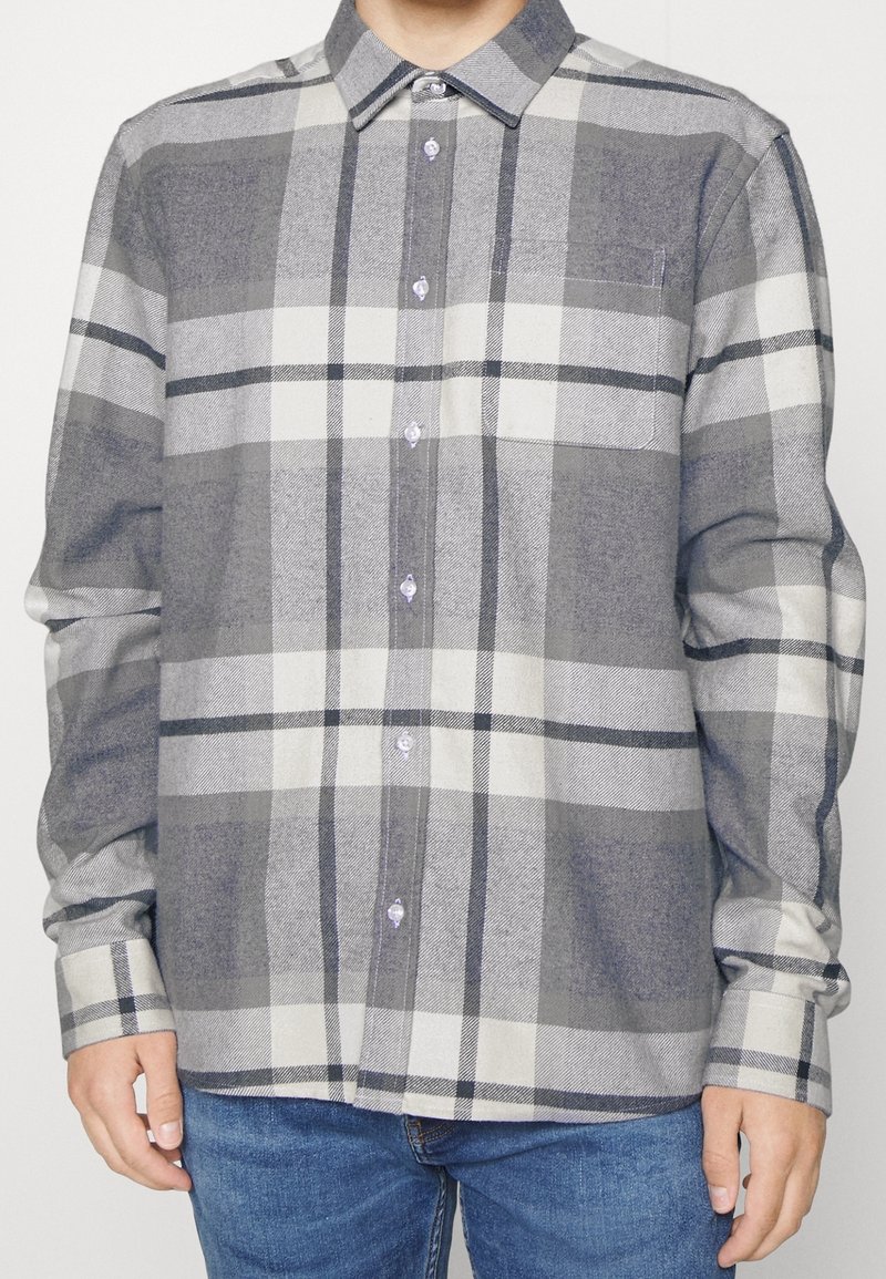 Homme portant une chemise à grands carreaux grise et blanche, boutonnée, à manches longues, et un jean bleu, debout devant un fond uni.