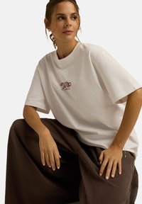 Biała koszulka z bawełny z haftem w kolorze bordowym przedstawiającym niedźwiedzia na klatce piersiowej, zestawiona z luźnymi brązowymi spodniami. Luzny krój i krótkie rękawy.