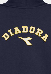 Jūras zilas džemperis ar izliekta dzeltena "DIADORA" teksta un baltu logotipa akcentu. Gluda tekstūra ar klasisku sporta dizainu.