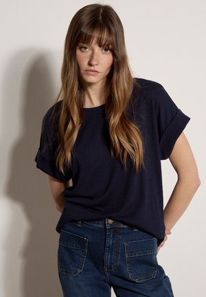 Femme aux longs cheveux bruns portant un haut à manches courtes bleu marine et un jean bleu, debout les bras derrière le dos devant un fond uni.