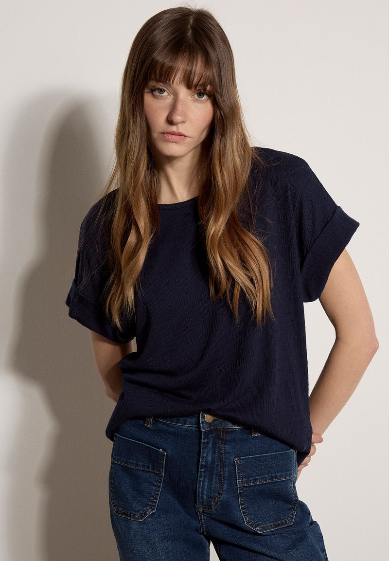 Frau mit langen braunen Haaren, die ein marineblaues Kurzarmshirt und blaue Jeans trägt, mit den Armen hinter dem Rücken stehend vor einem einfarbigen Hintergrund.