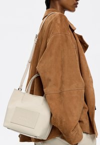 Borsa a spalla in pelle beige con finitura liscia, forma strutturata e manici superiori. Presenta un logo in rilievo sulla parte anteriore e una tracolla regolabile.