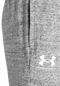 Pelēki sporta bikses, kas izgatavotas no mīkstas, teksturētas auduma, ar sānu kabatām un baltu Under Armour logo priekšpusē.
