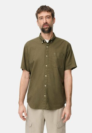 Mand med skæg iført olivengrøn kortærmet skjorte med knapper og beige cargobukser, stående mod en ensartet lys baggrund.