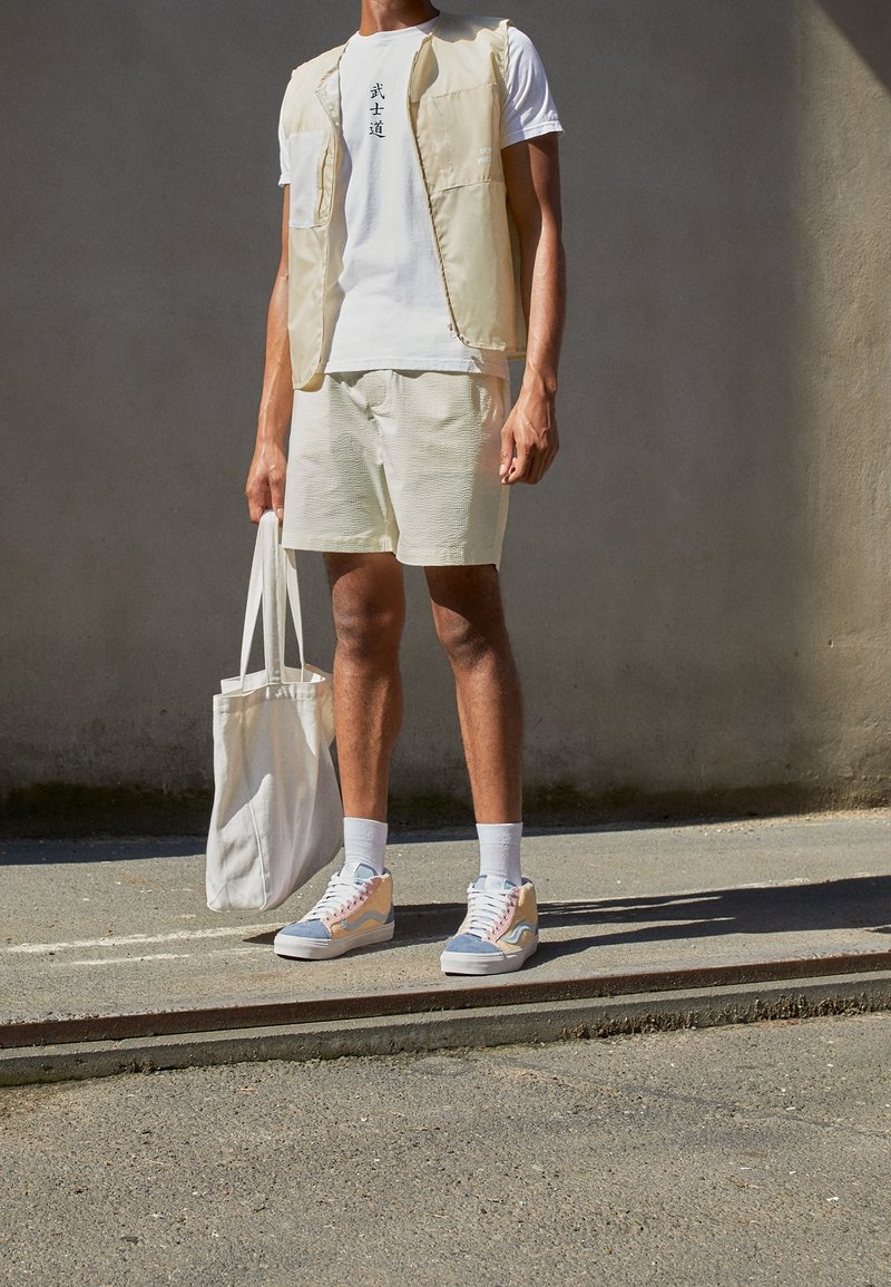 Homme portant un gilet beige, un t-shirt blanc avec un texte noir, un short beige, des baskets colorées, tenant un sac fourre-tout blanc, debout sur le trottoir.
