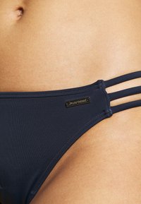 Navy bikini bottom with three strap accents on each side, made of smooth fabric, featuring a small metallic logo tag.