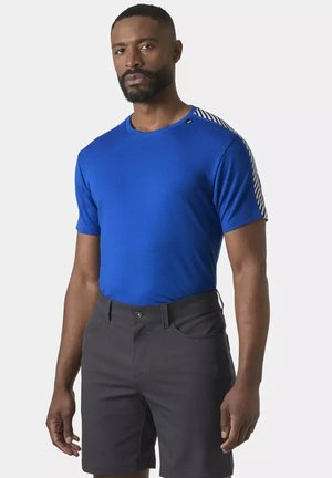 Uomo che indossa una maglietta blu a maniche corte con dettagli a righe sulle spalle e pantaloncini grigio scuro su sfondo semplice.