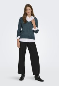 Türkisfarbener Strickpullover mit V-Ausschnitt, langen Ärmeln und geripptem Saum, über einem weißen Hemd mit Kragen getragen. Kombiniert mit schwarzen weiten Hosen und Stiefeletten.