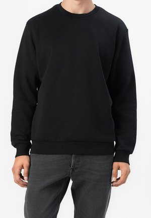 Hombre vistiendo una sudadera negra de manga larga lisa y jeans gris oscuro, de pie contra un fondo blanco.