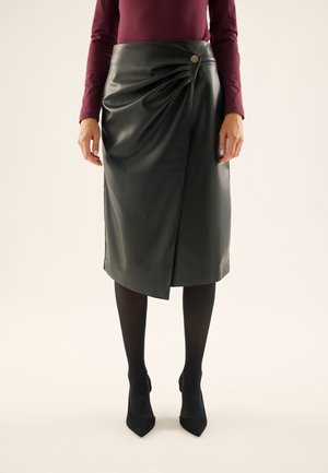 Femme portant une jupe portefeuille en similicuir noire avec un bouton sur le côté, des collants noirs, des talons noirs et un haut bordeaux à manches longues.