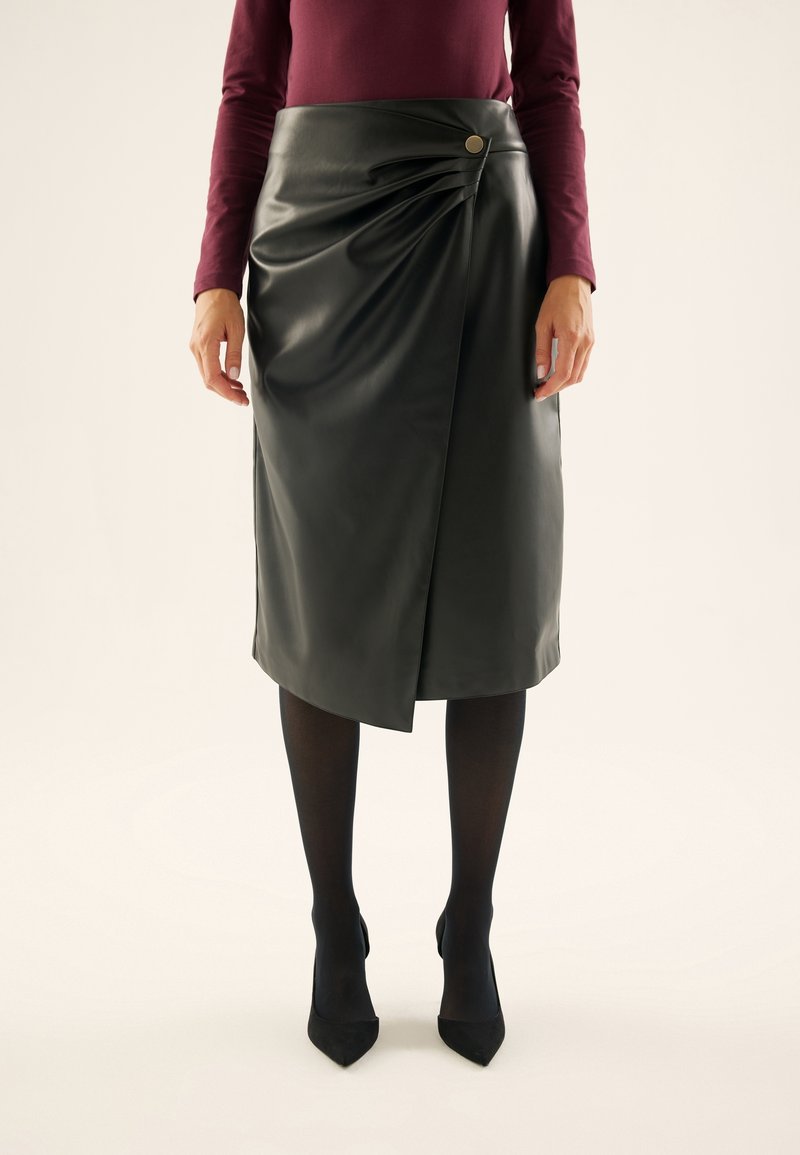 Femme portant une jupe portefeuille en similicuir noire avec un bouton sur le côté, des collants noirs, des talons noirs et un haut bordeaux à manches longues.