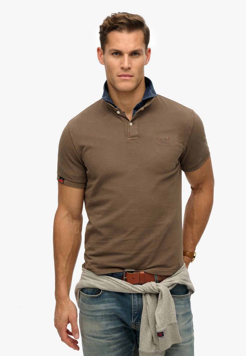 Polo shirt marrone a maniche corte con colletto in denim, logo ricamato sul petto, abbinato a jeans azzurri e un maglione grigio legato in vita.