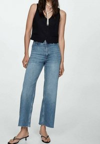 Chaleco negro sin mangas con botones, combinado con vaqueros acampanados azul claro. Los vaqueros tienen dobladillos sin rematar y un diseño ligeramente de talle alto. Sandalias negras.