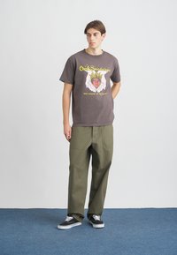 Γκρι γραφικό μπλουζάκι με κίτρινο και ροζ σχέδιο, συνδυασμένο με χακί παντελόνι cargo και μαύρα αθλητικά παπούτσια σε μπλε δάπεδο.