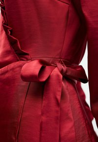 Robe rouge avec une finition satinée, ornée de volants texturés sur le côté et d'un nœud noué à la taille, mettant en valeur une silhouette lisse.