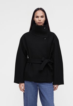 Une femme porte un manteau noir ceinturé avec un col montant et un jean bleu sur un fond clair uni.