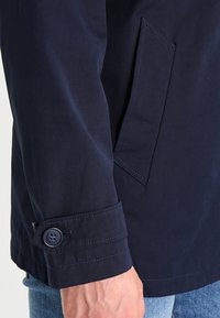 Marineblå trenchcoat i en glat, vævet stof. Har en sidelomme, manchet med knap og detaljerede sømme samt en skræddersyet pasform.