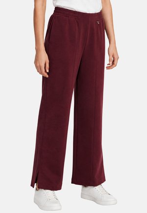 Donna che indossa pantaloni ampi color bordeaux con cuciture frontali e piccole aperture alle caviglie, abbinati a sneakers bianche e top bianco.