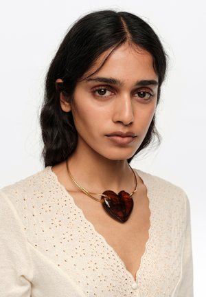 Mujer de cabello oscuro que lleva una blusa beige con ojales y un collar dorado con un gran colgante en forma de corazón marrón oscuro, mirando directamente al frente.