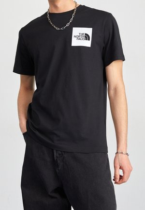 Black cotton t-shirt featuring a white logo patch on the chest. Short sleeves, round neck, and a relaxed fit. Worn with dark denim pants.