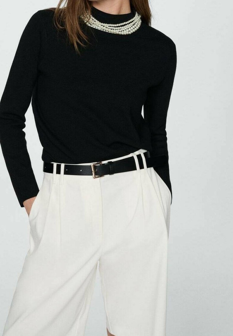 Femme portant un haut noir à manches longues, un collier de perles superposé, un short blanc taille haute et une ceinture noire avec une boucle dorée.