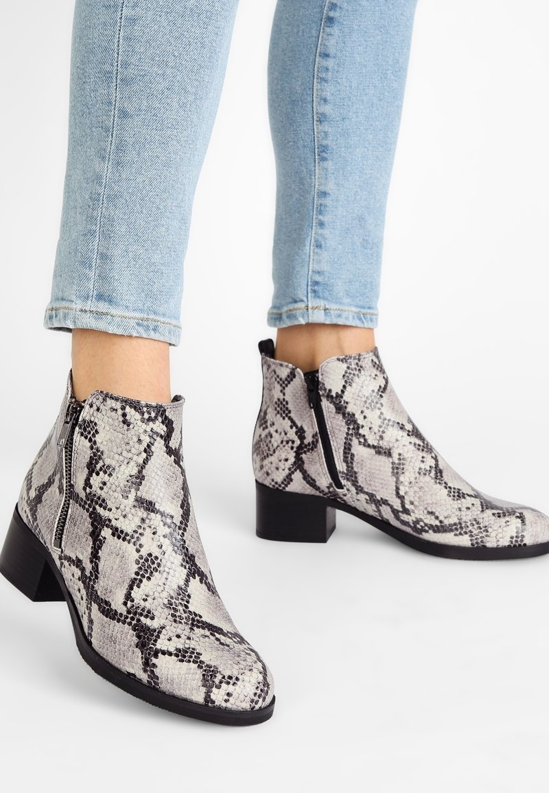 Las botas de tobillo de Python presentan una superficie texturizada, cremallera lateral y un tacón bajo. La paleta de colores incluye tonos grises y negros.