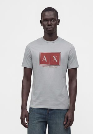 Ung mand med mørk hud iført en lysegrå Armani Exchange T-shirt og blå jeans mod en ensfarvet hvid baggrund.