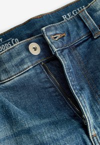 Modré džínsy z denimu s otvorením na zips, bronsovým gombíkom a kontrastným oranžovým prešívaním. Jemne opotrebovaný vzhľad, pravidelný strih.