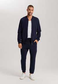 Chaqueta bomber navy con cremallera frontal, puños acanalados y joggers a juego, que presentan una cintura con cordón y un tejido suave. Zapatillas blancas complementan el conjunto.