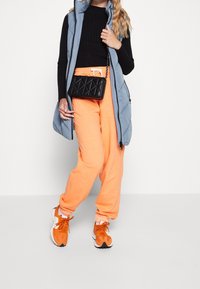 Person in orange Jogginghose, schwarzem geripptem Pullover, blauer ärmelloser Steppweste, orangefarbenen Sneakers und trägt eine schwarze gesteppte Umhängetasche.
