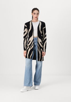 Long cardigan avec un motif rayé zèbre noir et crème, design à front ouvert, texture en maille, associé à un jean large bleu clair et des chaussures blanches.