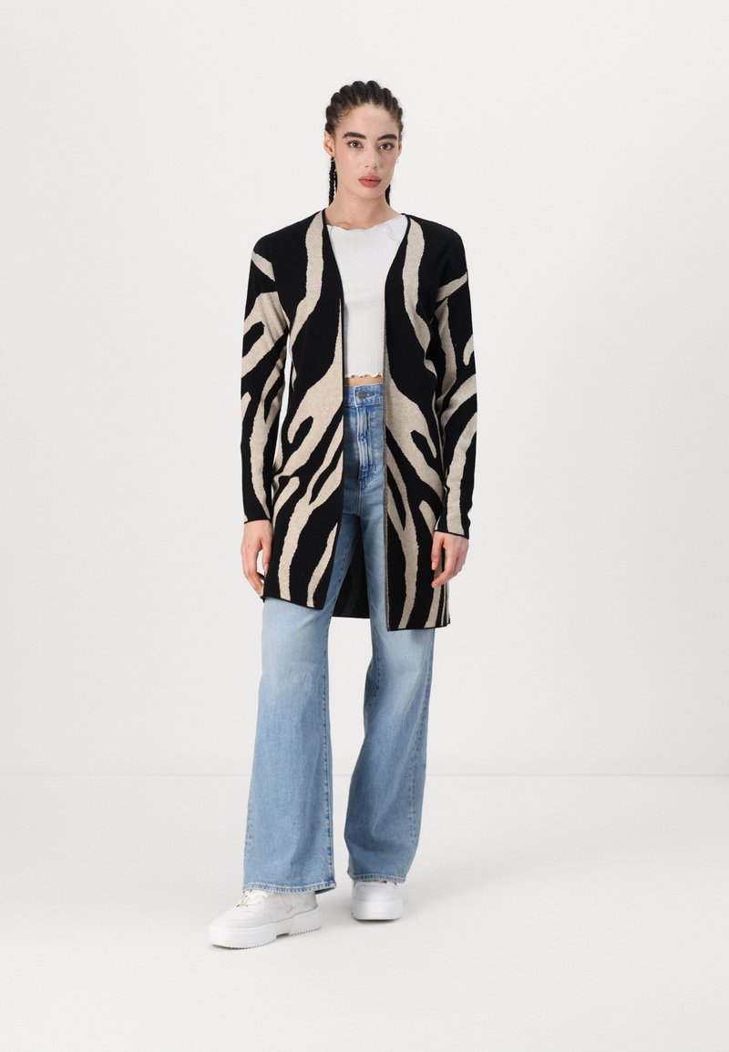 Garšs kardigāns ar melnbaltu zebra svītru rakstu, atvērta priekšpuse, adīta tekstūra, kombinēts ar gaiši ziliem platām kājām džinsiem un baltām kurpēm.