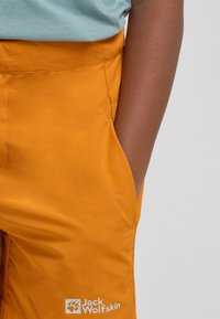 Orangene Shorts aus leichtem Material, ausgestattet mit einer Seitentasche und einem Logo am unteren Bein. Die Textur scheint glatt und langlebig zu sein.