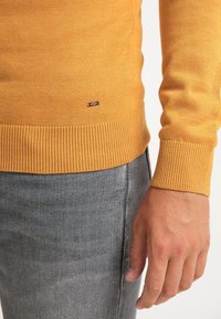 Senapsgul tröja i stickad tyg, med ribbade muddar och nederkant, samt en liten märkeslapp på nedre vänstra sidan. Buren över grå jeans.