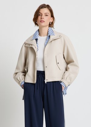 Donna che indossa una giacca corta beige con polsini elasticizzati sopra una camicia a righe blu e un top bianco a coste, abbinata a pantaloni blu navy con coulisse.