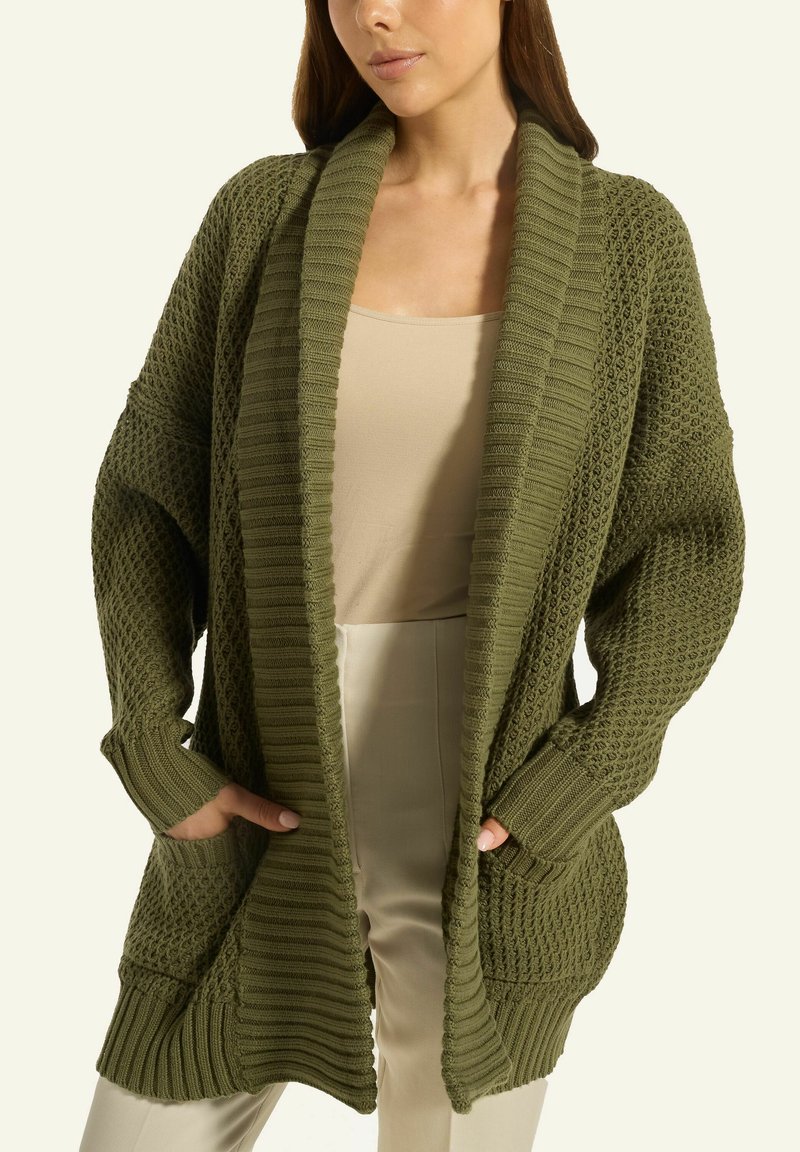 Frau, die eine übergroße olivgrüne Strickjacke über einem beigen Oberteil und einer Hose trägt, mit den Händen in den Taschen der Strickjacke, steht vor einem einfarbigen Hintergrund.