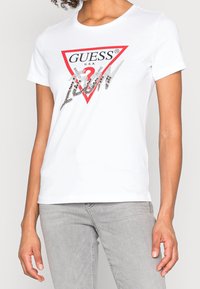 Hvid kortærmet t-shirt med et rødt trekantet logo, der har "GUESS" og "1981" i sølvdetaljer og en glat bomuldsstruktur.