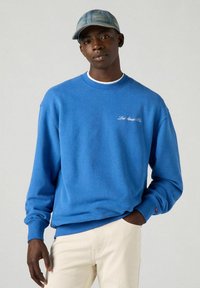 Sudadera azul con cuello redondo, puños y dobladillo de canalé. Presenta texto bordado en blanco. Se lleva con pantalones de color claro y una gorra de mezclilla.