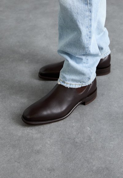 Braune Lederstiefel mit glatter Textur, spitzer Zehenform und brauner Gummisohle, kombiniert mit hellblauen Jeans.