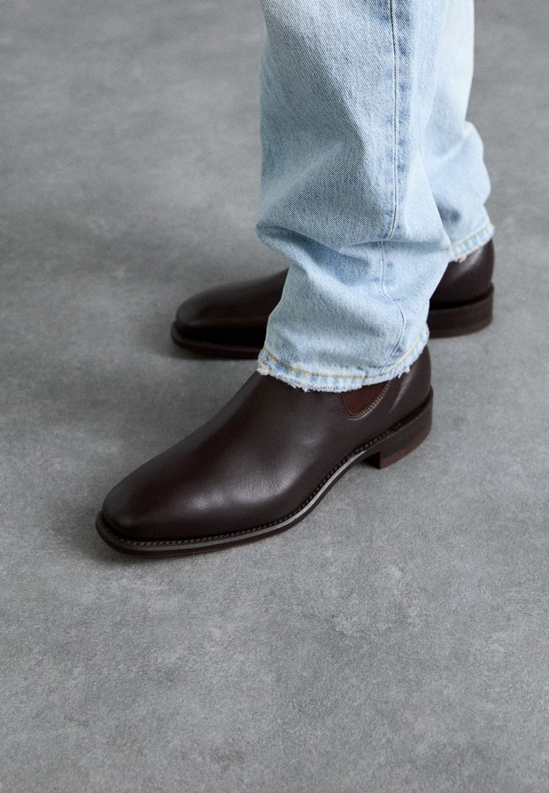 Pruuni nahast ankle boots, millel on sile tekstuur, terav nina ja pruuni kummitald, koos helesiniste denim teksadega.