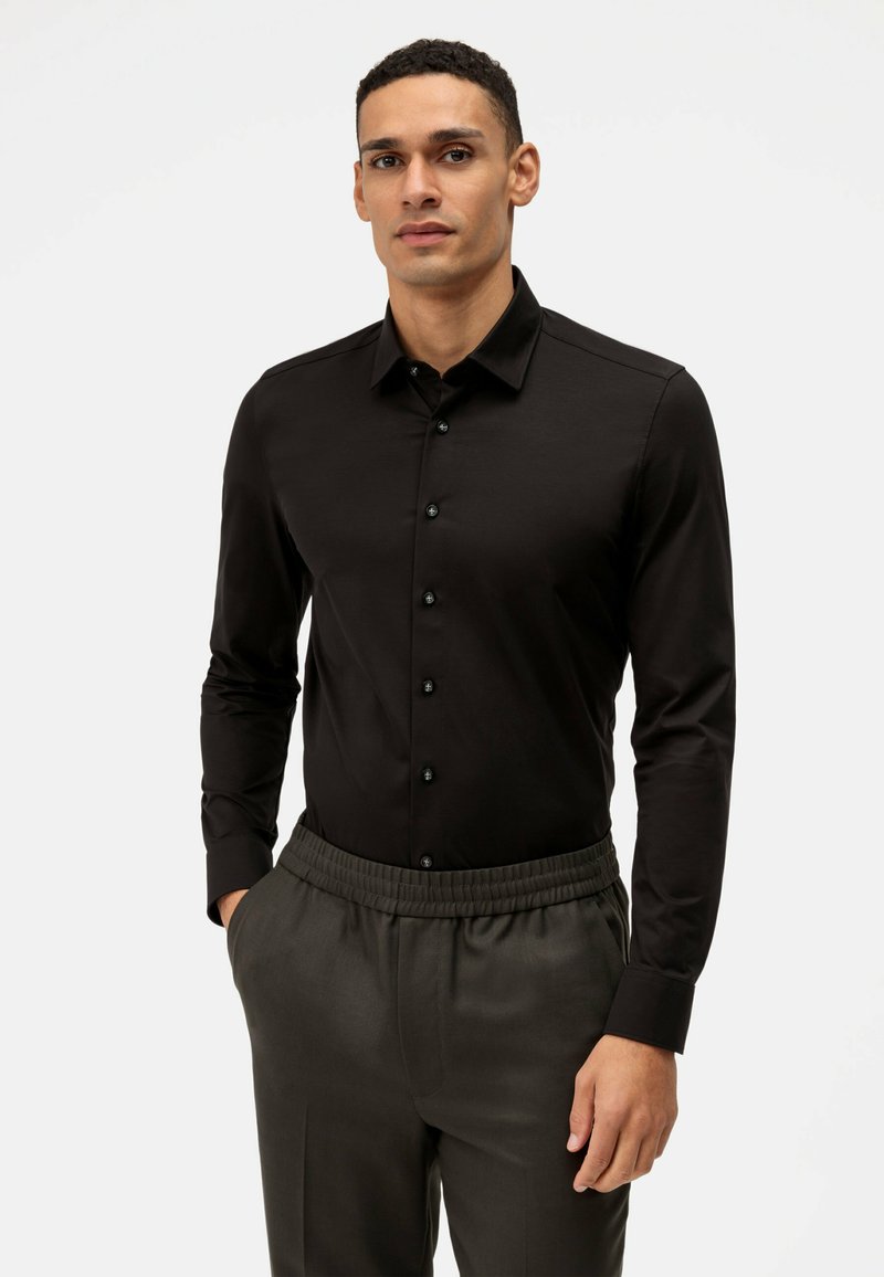 Zwarte longsleeve shirt met een kraag, voorzien van zwarte knopen en gladde stof; gecombineerd met grijze broek met elastische tailleband.