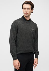 Νεαρός άνδρας με σκούρο γκρι πουλόβερ με στρογγυλή λαιμόκοψη πάνω από ένα μαύρο πουκάμισο με γιακά, που στέκεται μπροστά σε ένα απλό ανοιχτό φόντο.